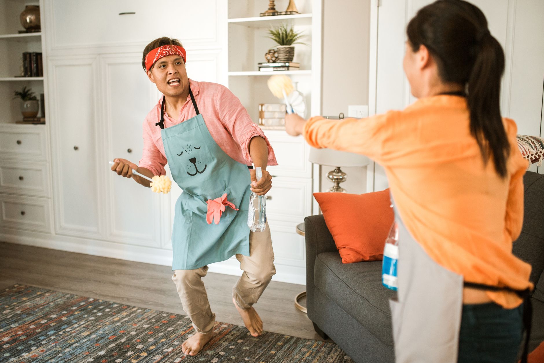 Two people playfully cleaning indoors, one wearing a blue apron with a bear design and a red bandana, holding a scrubber and a spray bottle, while the other, in an orange shirt, reaches out with another cleaning tool.