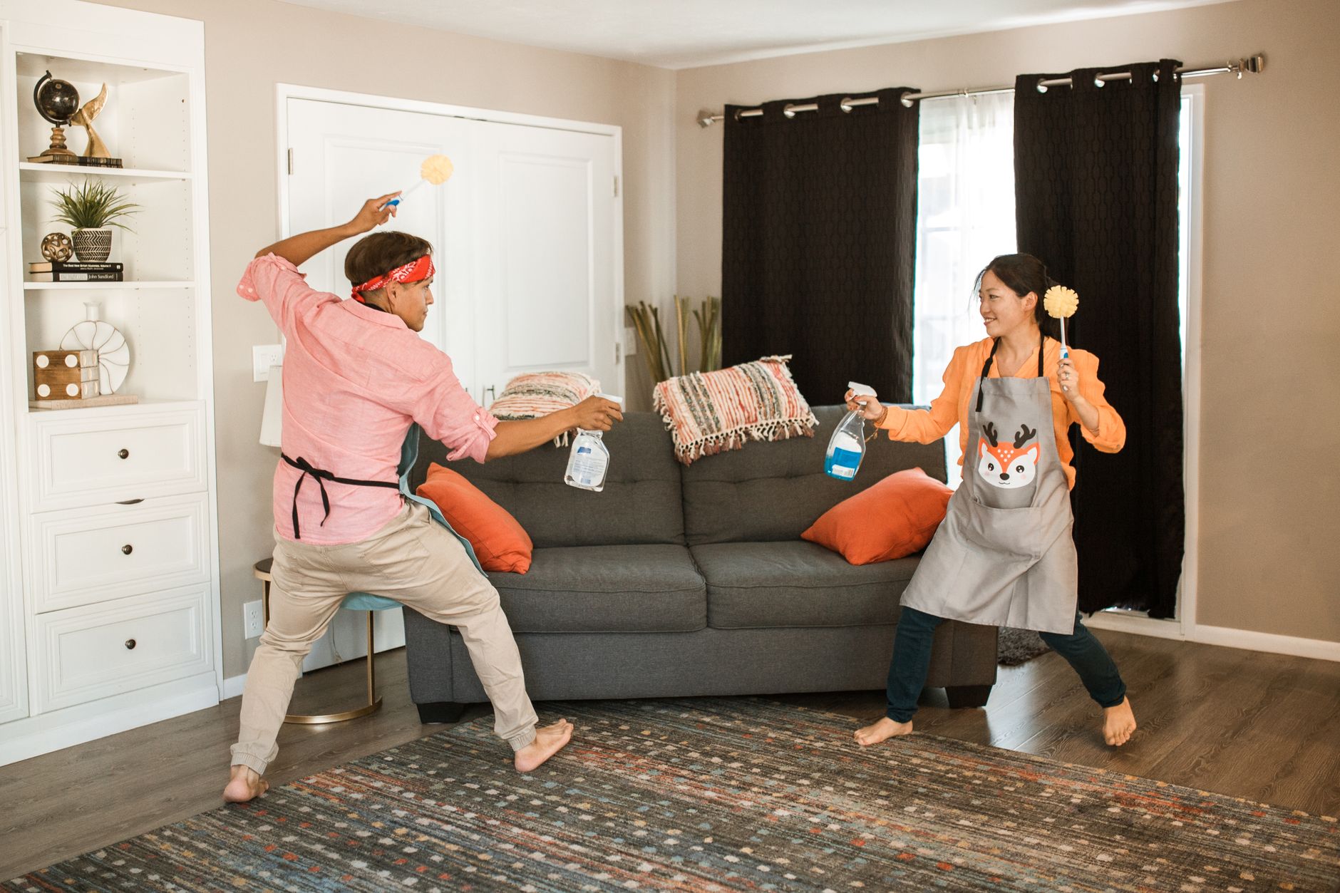 A man and a woman playfully cleaning a living room. The man, wearing a pink shirt and a bandana, holds a cleaning spray, while the woman, in an orange shirt and an apron with a fox design, wields a cleaning tool. They appear to be having fun in a casual and bright interior setting.