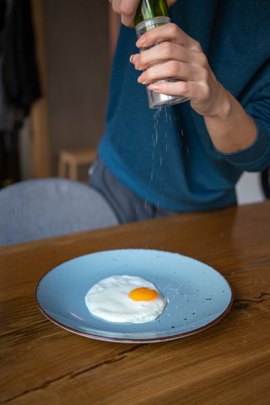 A close-up of a hand sprinkling salt over a fried egg on a light blue plate, with a wooden table in the background.