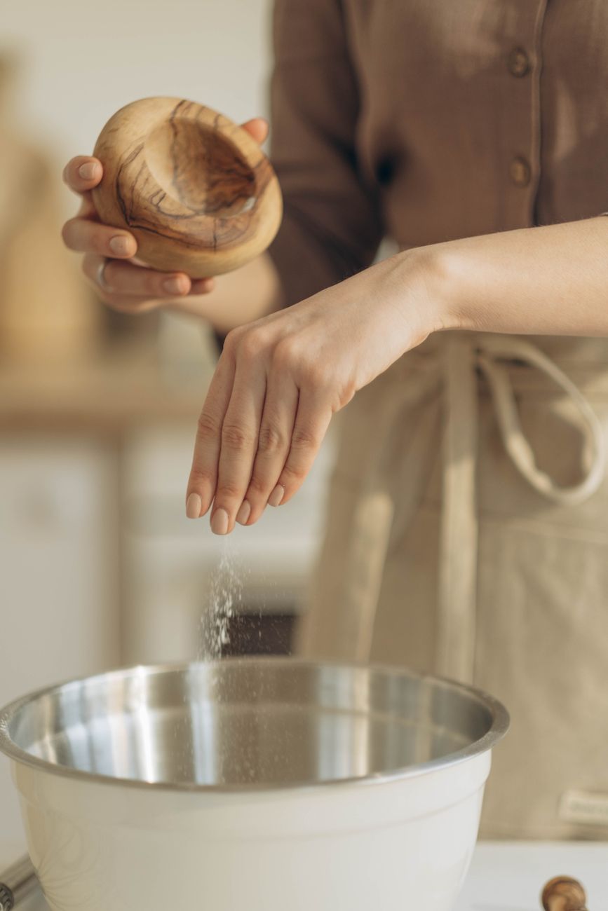 A person in an apron sprinkles salt from a wooden bowl into a metal mixing bowl in a bright kitchen.